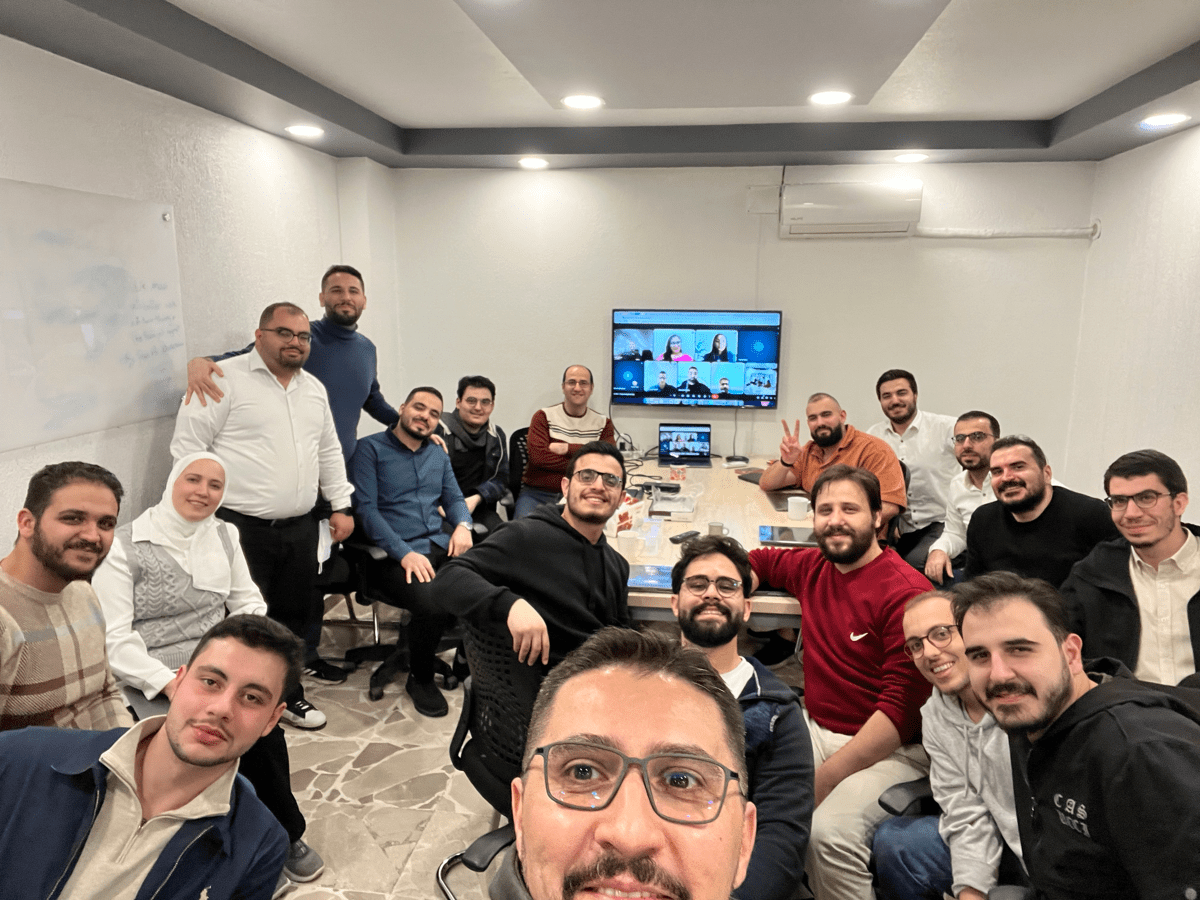 Group selfie in a conference room during a hybrid meeting, with colleagues around a table and a video call displayed on a screen.
