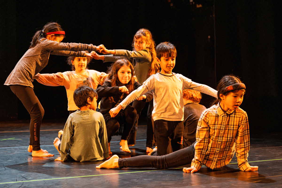 Children dancing together on a dark stage, using their arms to make shapes and connect themselves to one another