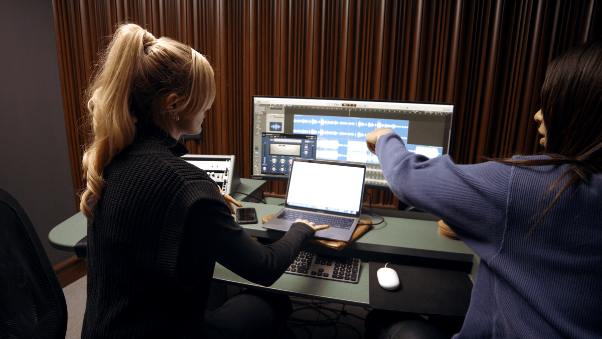An employee and an artist sitting in the music studio looking at a screen which shows the sound waves of a track that they are pointing to and discussing 