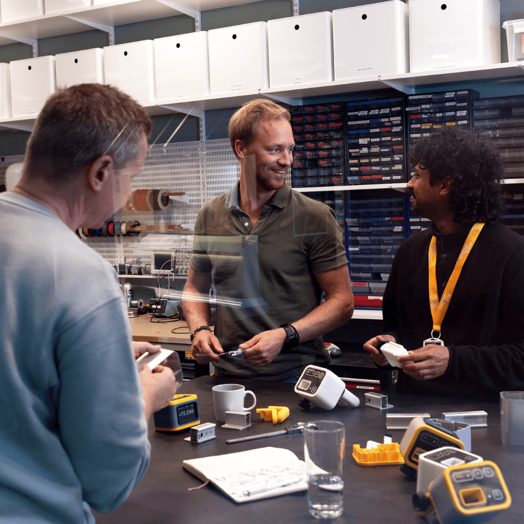 Three men standing around a work table full of hardware