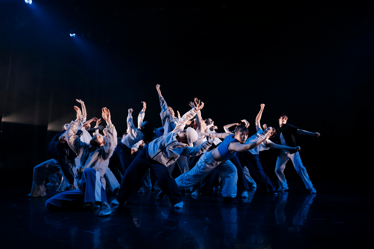 Image of a large group of dancers in white tracksuits under blue light on an otherwise empty stage.