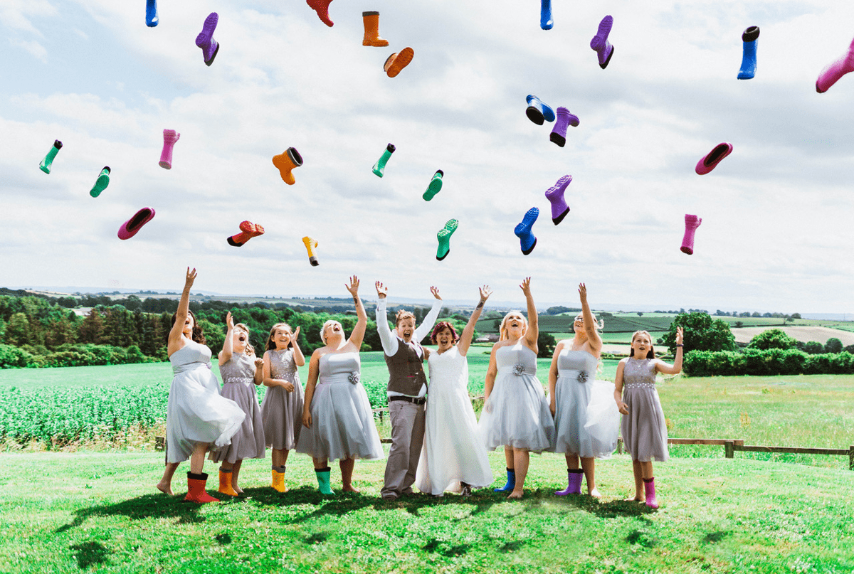 A newly-wed couple stand in a field with bridesmaids either side in dresses and one brightly-coloured welly each. Their other welly (and many more) have been thrown in the air to create a welly rainbow! Everyone is smiling and laughing. 