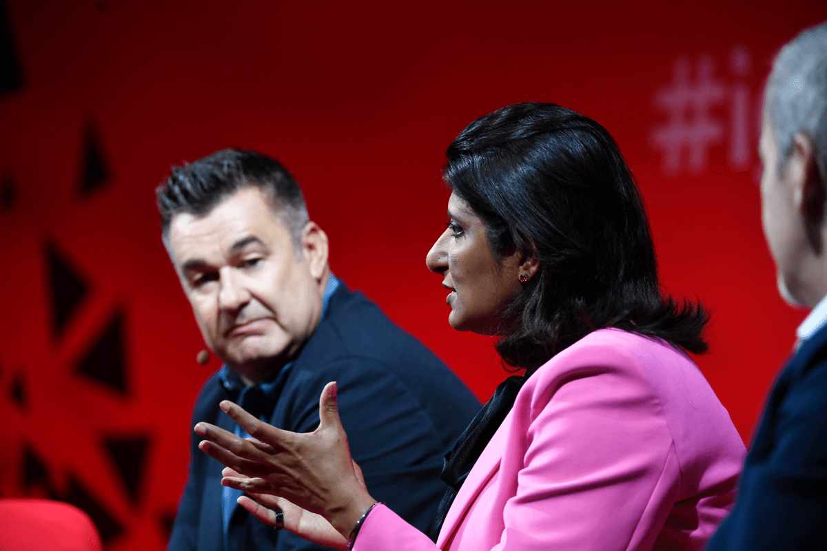 A close up shot of three TMF members sitting in an auditorium with a red background. One lady is side on and can be seen gesturing with her hands as her two colleagues are looking at her whilst they listen.