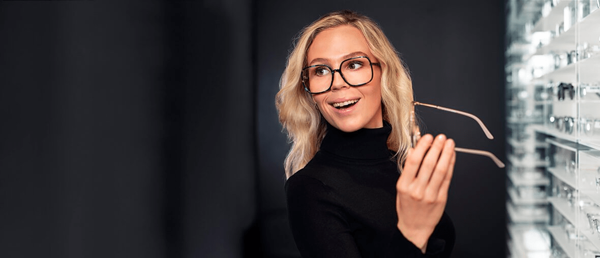 Hymyilevä nainen tummassa poolopaidassa kokeilee silmälaseja Synsam liikkeessä