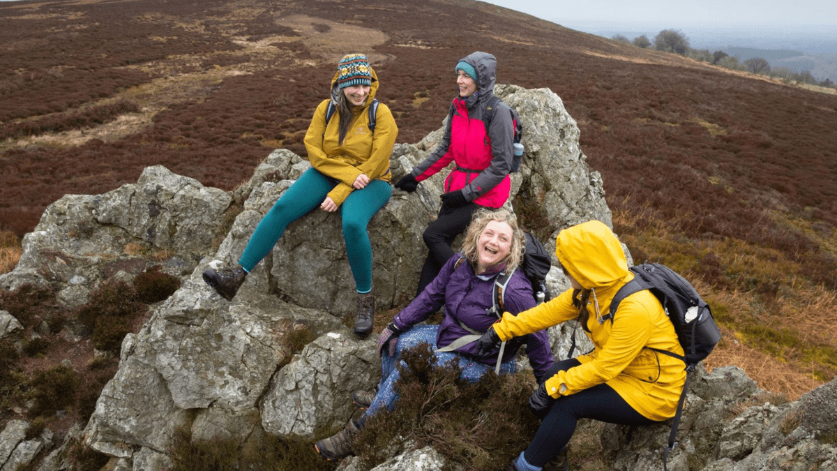 Four hikers in colourful waterproof jackets gather around rocks on a moorland hillside.