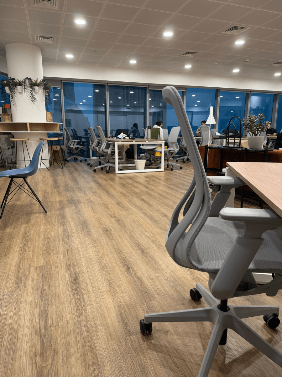 Modern open-plan office with ergonomic chairs and desks, glass wall meeting room, and warm ceiling lights.