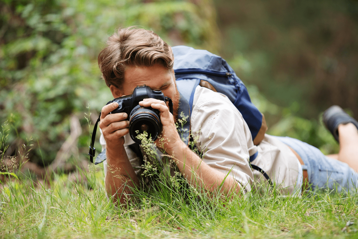 	En person ligger på marken i naturen och fotograferar med en kamera, bär ryggsäck.