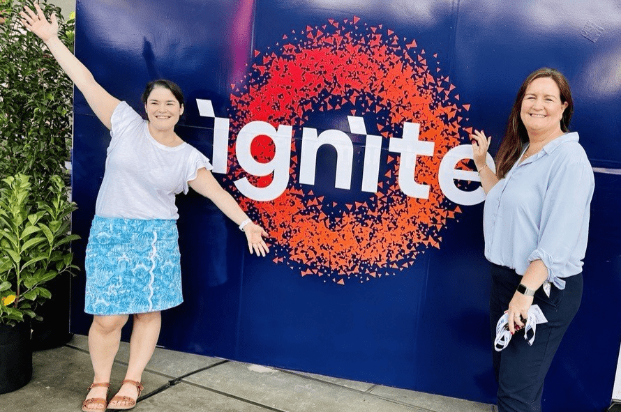 A background of the TM Forum Ignite logo. One female colleague is standing to the left with her arms open wide to highlight the sign, whilst a second colleague is side on to the camera with one hand raised touching the logo.