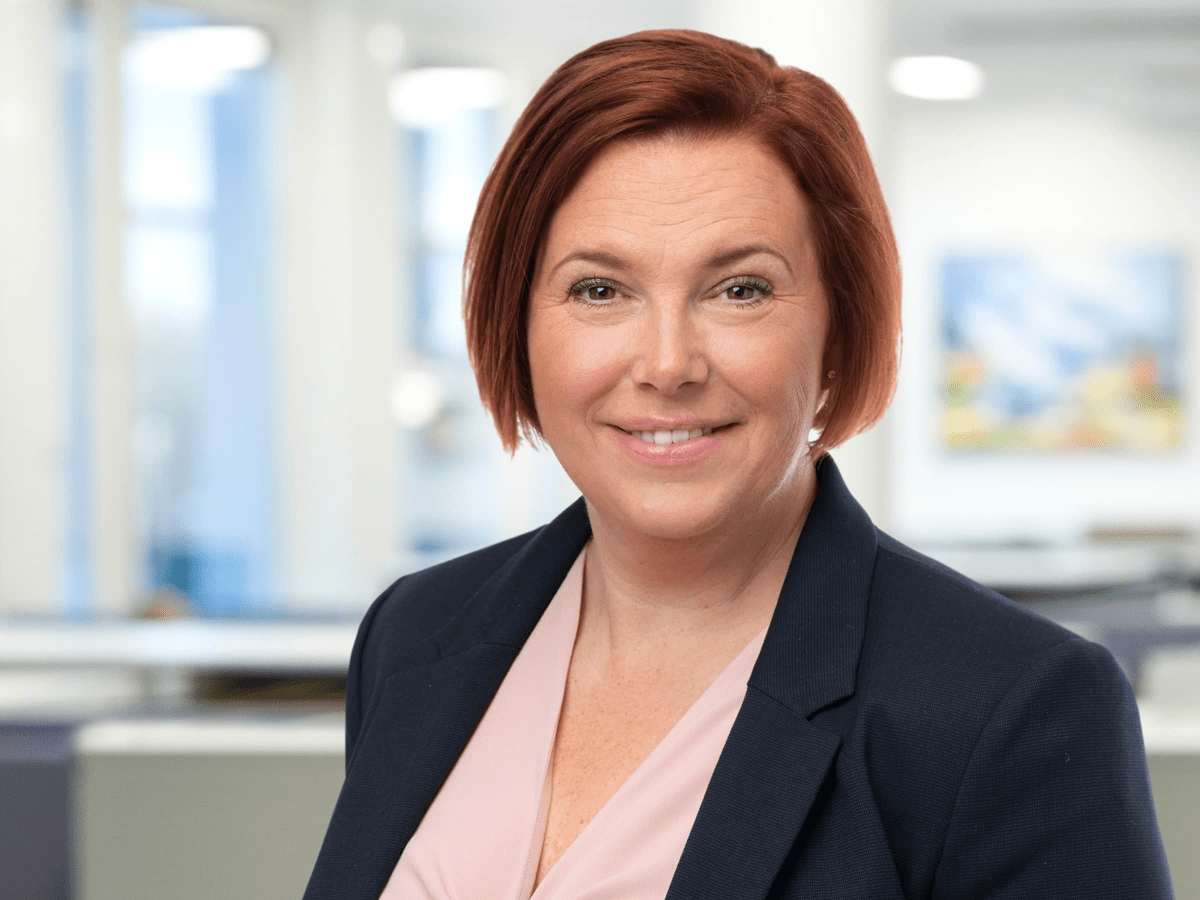 Kvinna i kavaj ler mot kameran i ljus kontorsmiljö med suddig bakgrund.