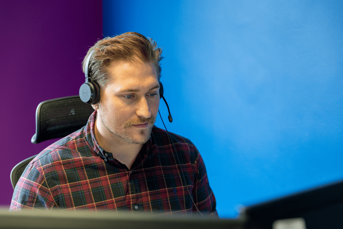 Man wearing a headset working at a computer against a blue and purple wall.