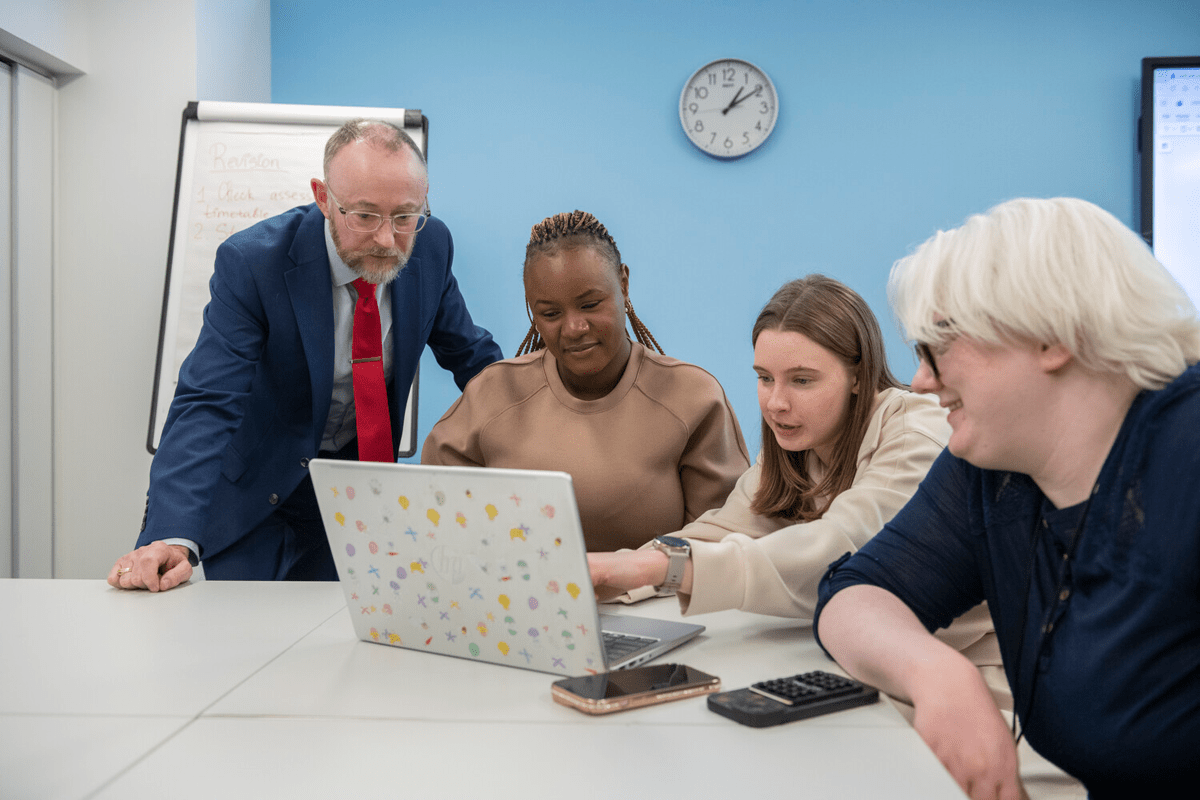 A man in a suit and red tie leans over a laptop, looking at the screen with three women.
