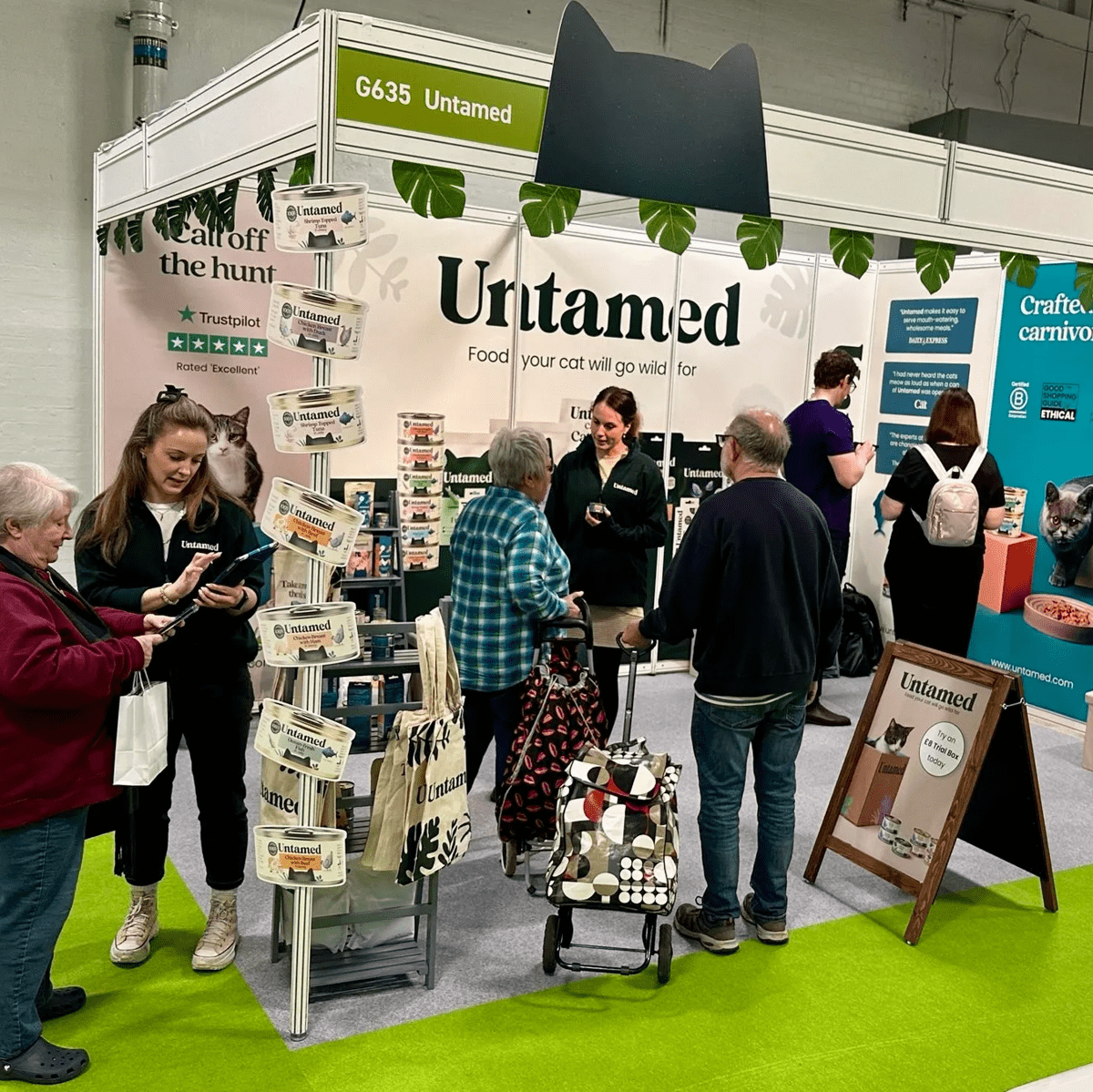 Customers and brand ambassadors look at Untamed products whilst standing in front of a large branded Untamed display at one of our events. Our tins, branding, and reviews are clearly displayed.
