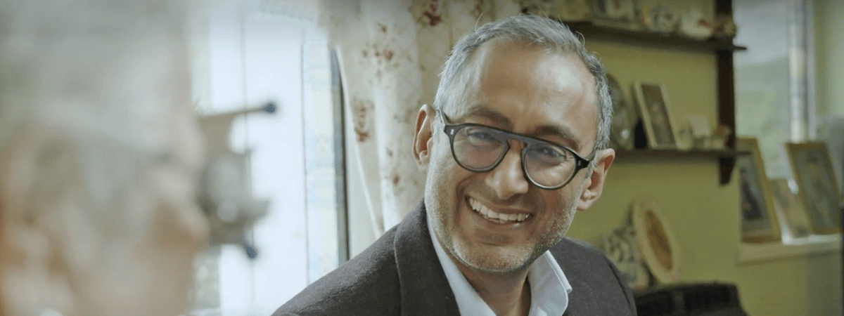 Mam, smartly dressed with glasses on smiling, he is talking to someone and behind him are curtains and shelves with photos on
