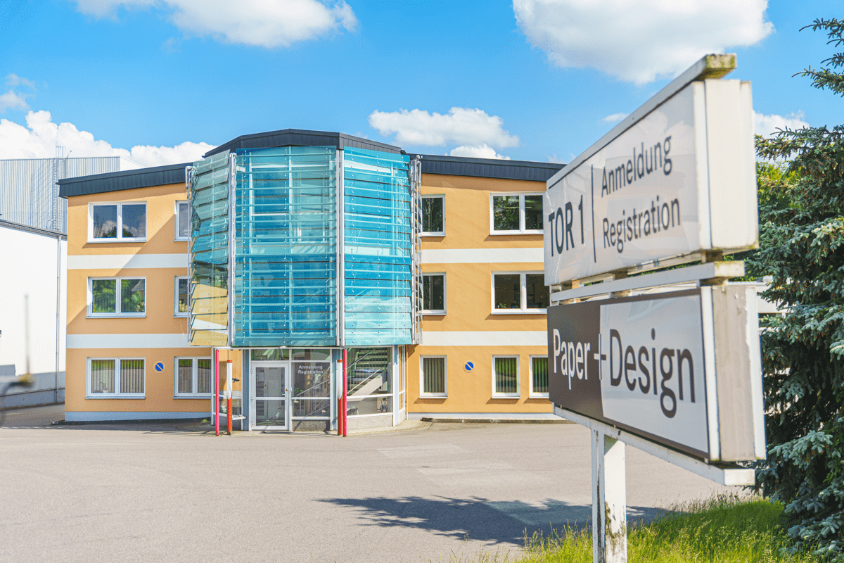 Eingangsbereich des Firmengebäudes von Paper + Design GmbH Tabletop mit moderner Glasfassade und Hinweisschild zur Anmeldung (Tor 1) bei sonnigem Wetter.