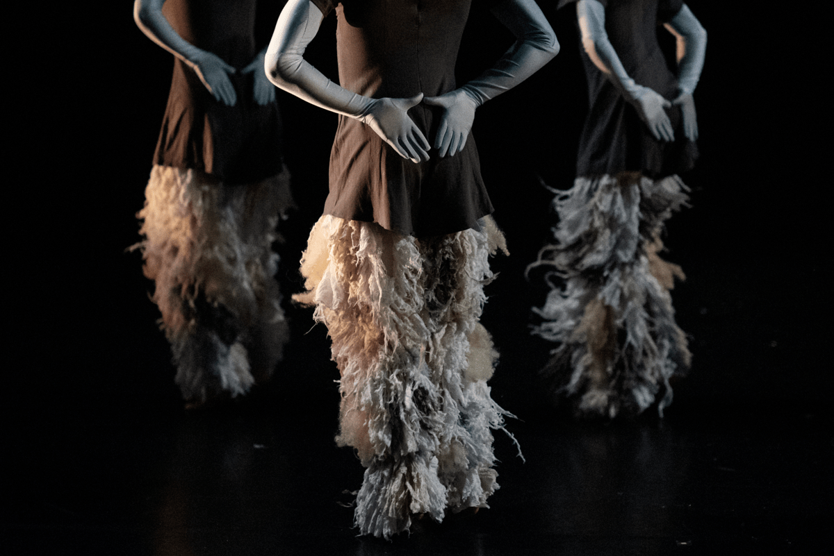 Image of three dancers from the shoulders down. They are facing with their backs to the camera and holding their hands in triangle positions behind their backs. They are wearing loose brown tops, cream feathered, floor-length skirts and long gloves that cover their entire arms.