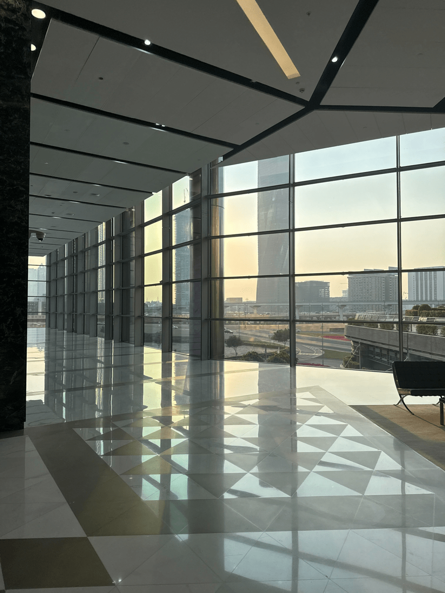 Spacious modern lobby with floor-to-ceiling glass windows, glossy patterned flooring, and a sunset city view outside.