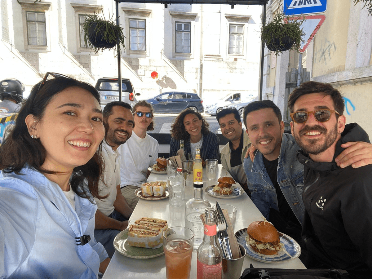 A group of coworkers sit together at a long outdoor café table, smiling at the camera while enjoying sandwiches, burgers, and drinks on a sunny street.