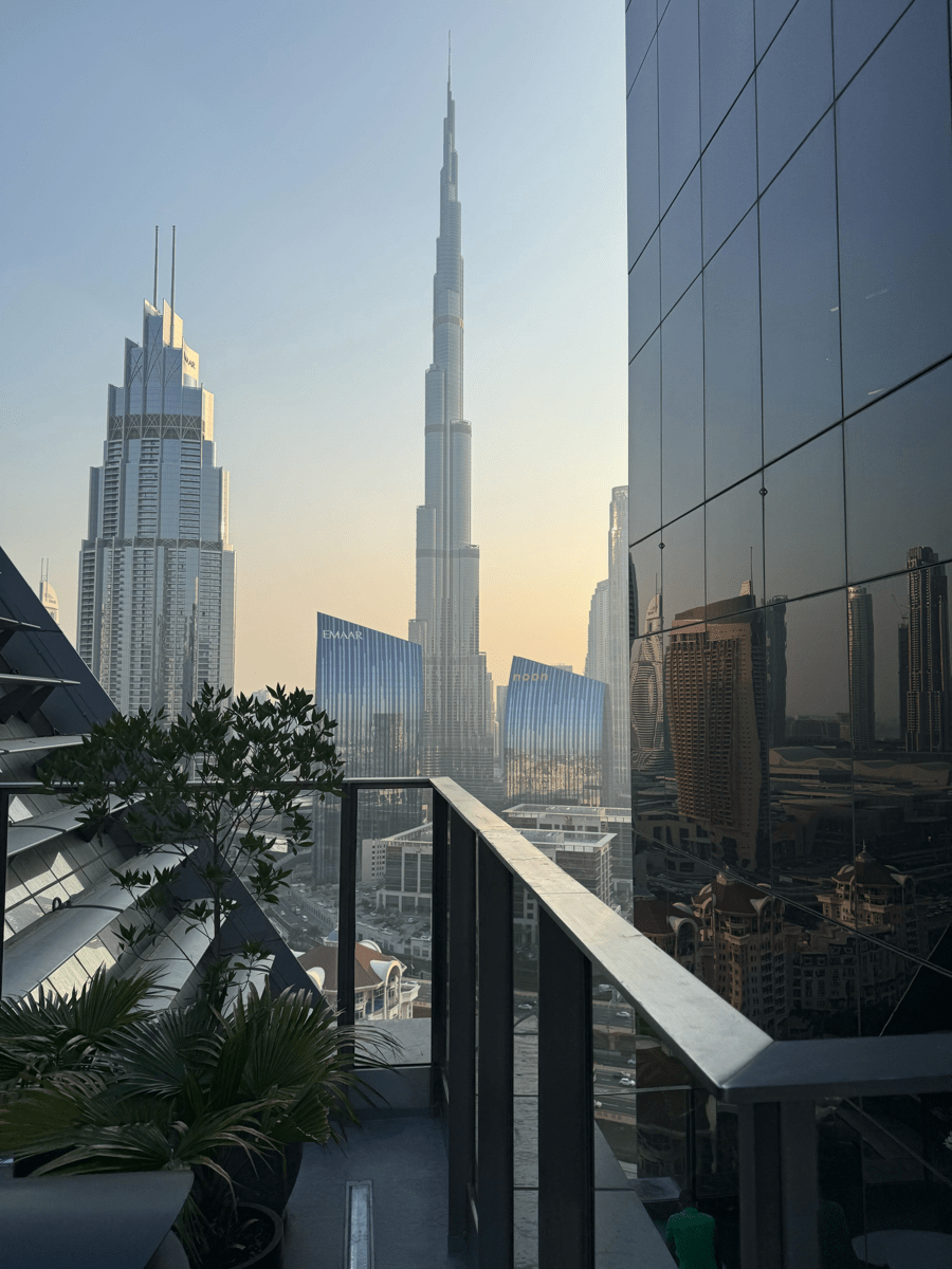 View from a terrace overlooking Downtown Dubai, with the Burj Khalifa centered and nearby towers reflected in a glass facade.