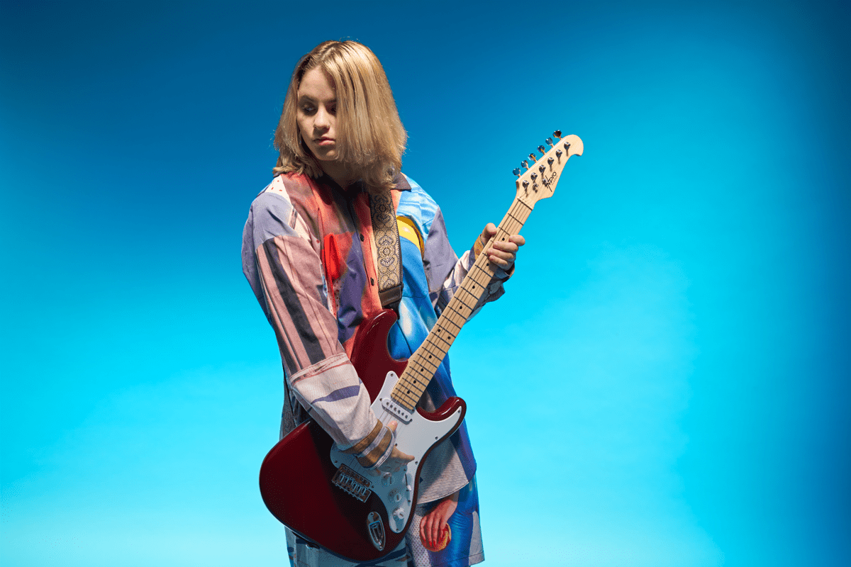 An artist holding a guitar and standing with a blue background behind them