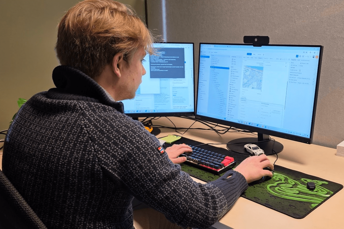 Utvikler hos Excito som sitter med ryggen til forran doble skjermer som viser no-code app-bygger. Han jobber på en bred gaming musmatte med mekanisk tastatur og en kul liten modellbil som bordpynt. 