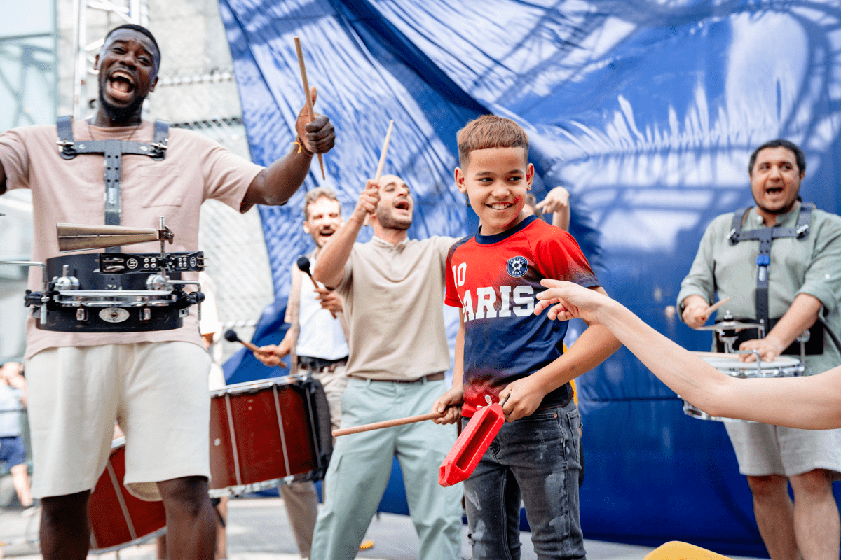 Performers stand drumming, alongside a child standing in the centre who is also trying out drumming with the group.