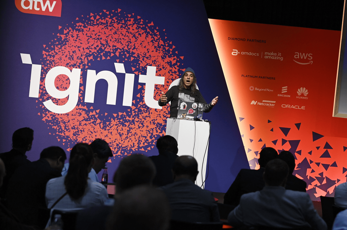 The DTW Ignite stage. A man dressed casually wearing a sweatshirt and beanie hat is on the stage talking and gesticulating with his hands. Before him you can see the darkened shadow of other TMF members who are listening to his talk.
