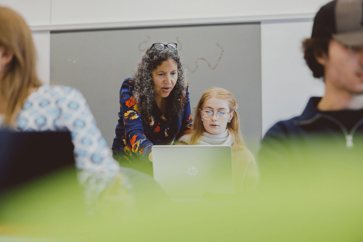 A woman in a floral shirt leans over a student wearing glasses and a tan sweater, both looking at a laptop in a classroom setting.