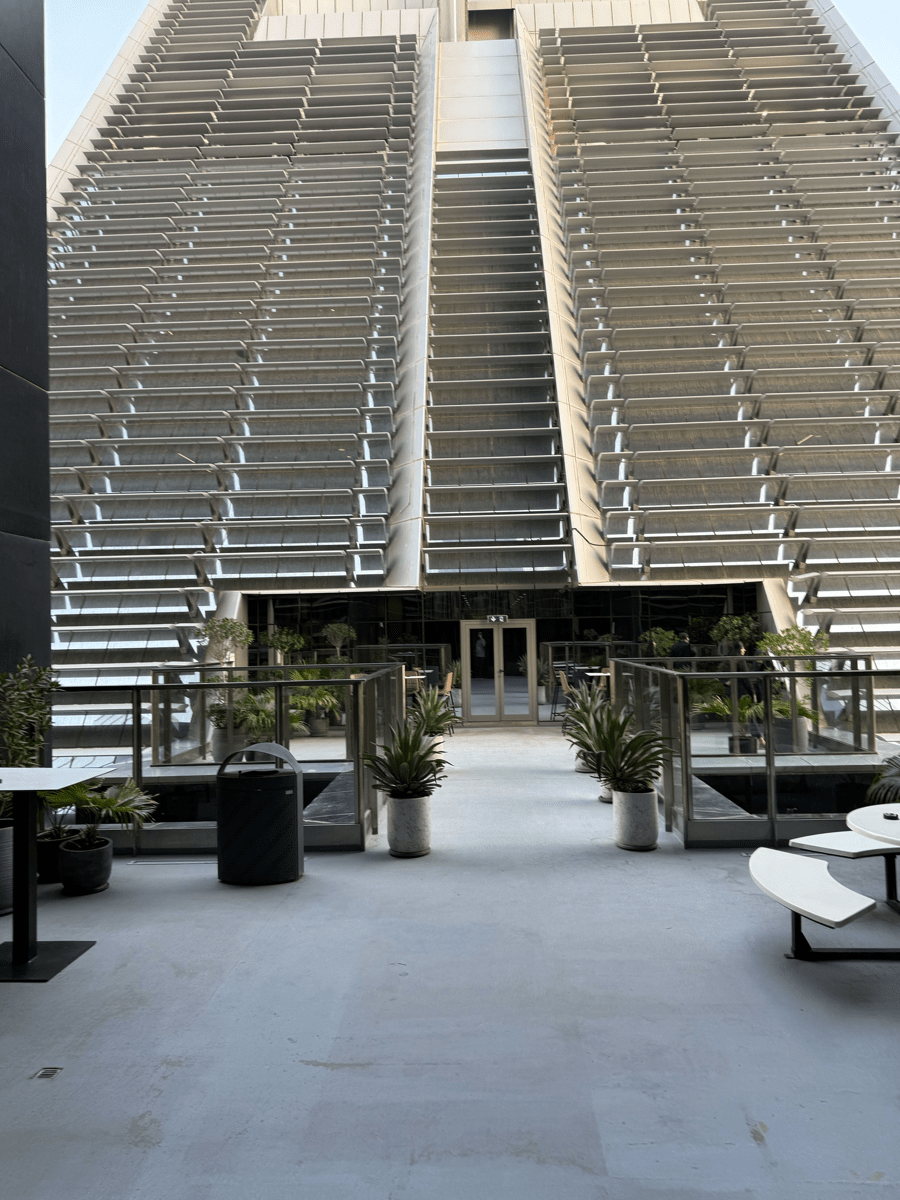 Outdoor walkway leading to a modern building entrance, framed by large louvered facades, glass railings, and potted plants.