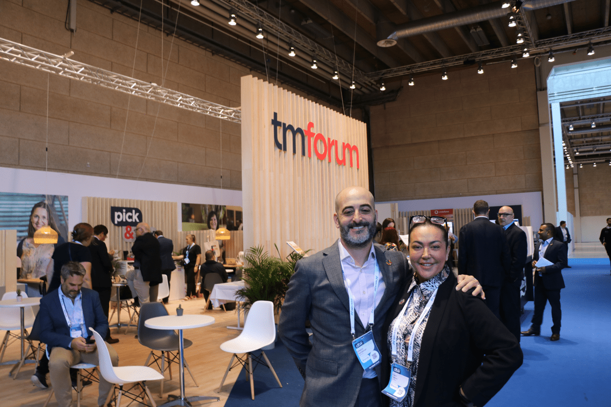 Two TMF colleagues standing to the right of the photo smiling at the camera. Behind them are other colleagues and members and a large wooden wall with the TM Forum written logo.