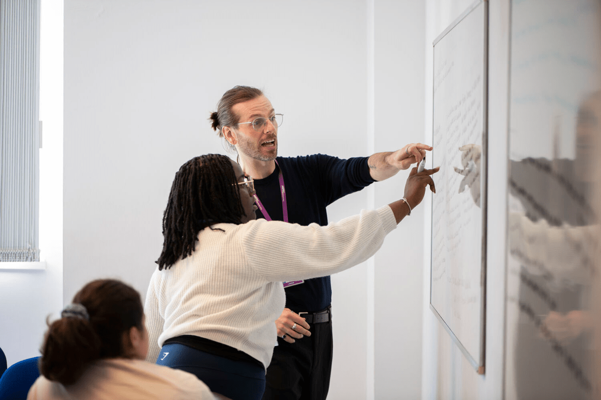 Two individuals, one with dark locs and wearing a white sweater, the other with brown hair pulled back and wearing a dark shirt and glasses, point at and interact with a whiteboard covered in handwritten text in a brightly lit room.