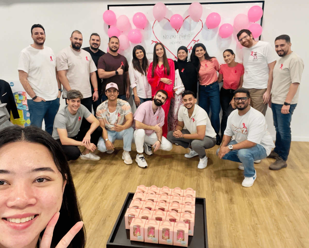 A diverse group of coworkers in casual clothes, many wearing pink and breast cancer ribbons, pose smiling in an office with pink balloons, a “No one fights alone” sign, and gift boxes, while a woman in front takes a selfie.