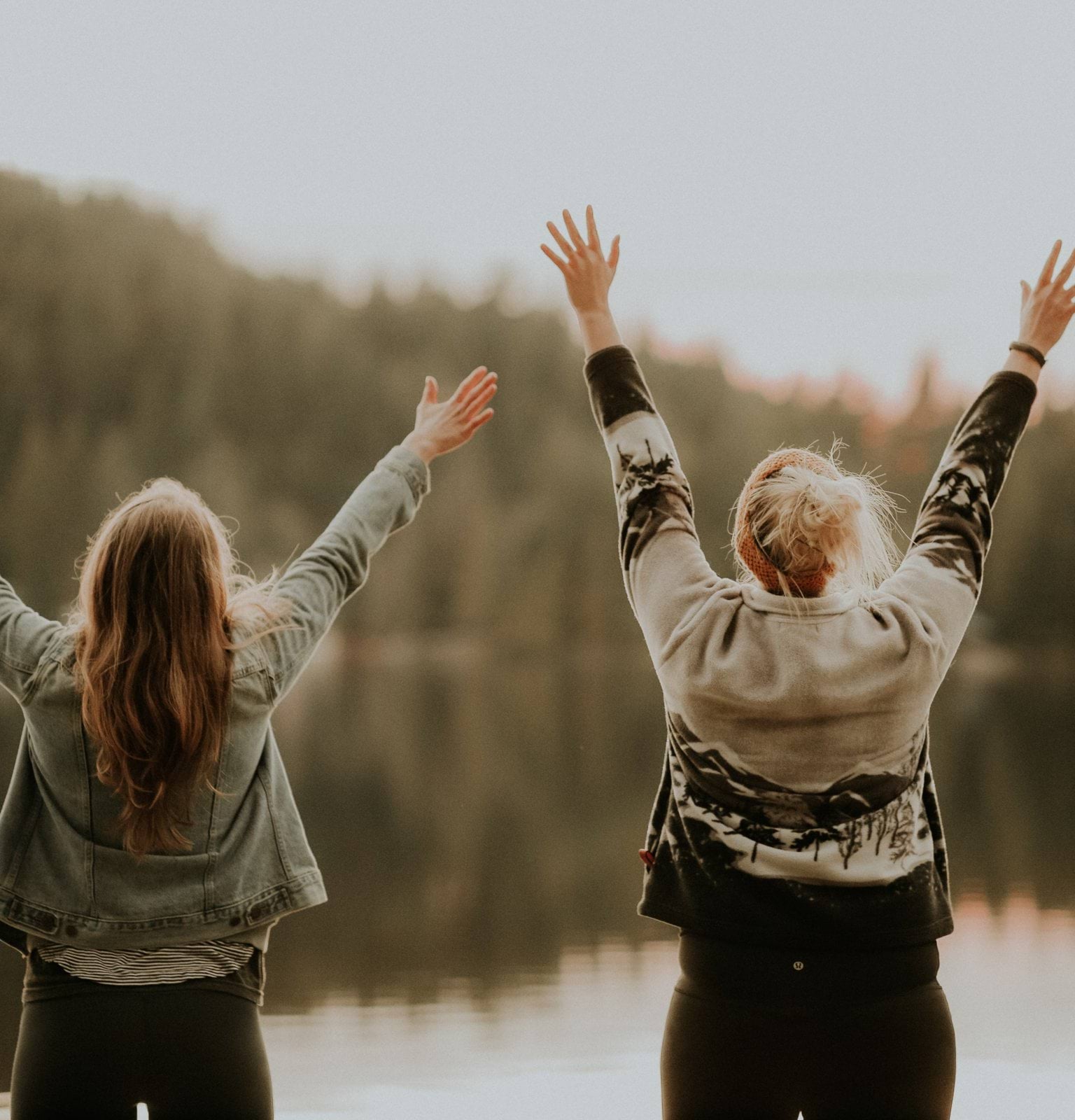 two women hands up standing beside body of water