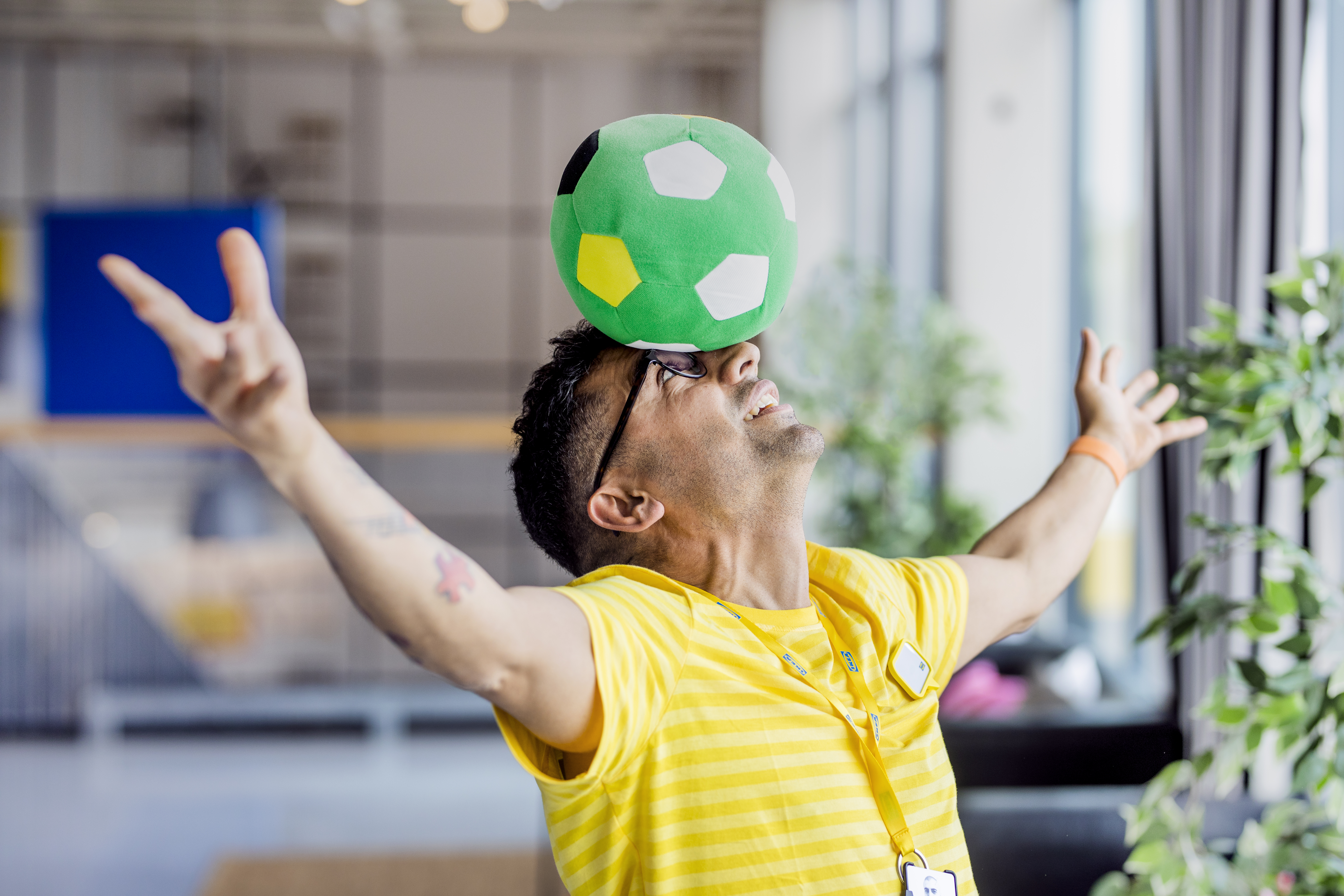 Geltoną uniformą vilkintis IKEA darbuotojas stovi ištiesęs rankas į šalis ir balansuoja ant galvos žalios spalvos žaislinį futbolo kamuolį.