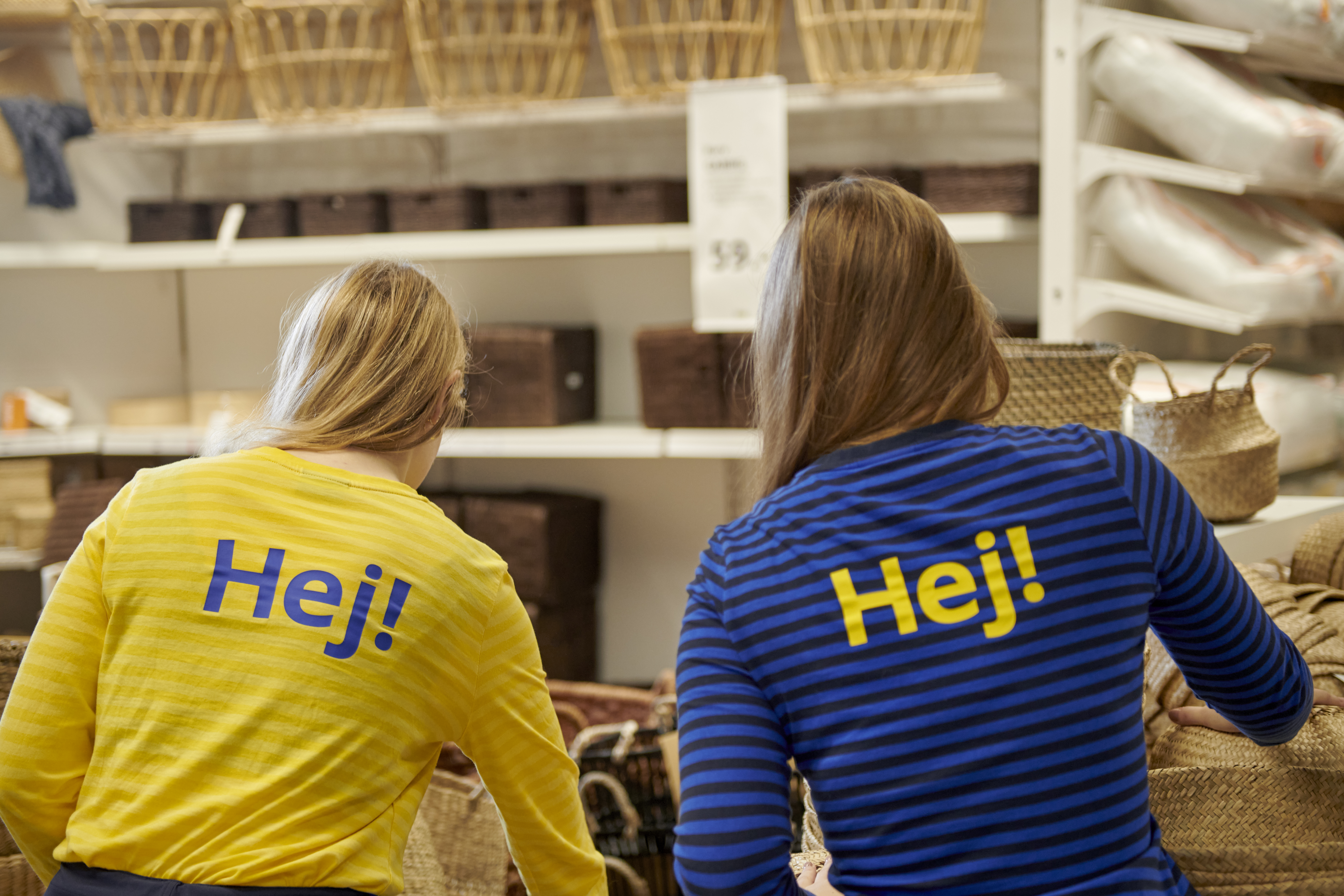 Kaks sinikollastes tööriietes IKEA töötajat selja tagant, kelle särkidel paistab sõna „Hej!“. Taustal on näha poodi koos korvide ja karpidega.