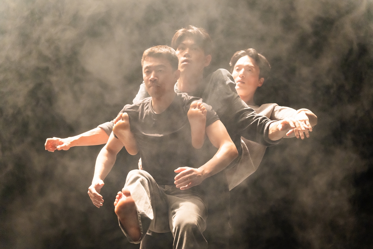 Image of 3 dancers on a stage, one carrying the other two in their back, surrounded by haze.