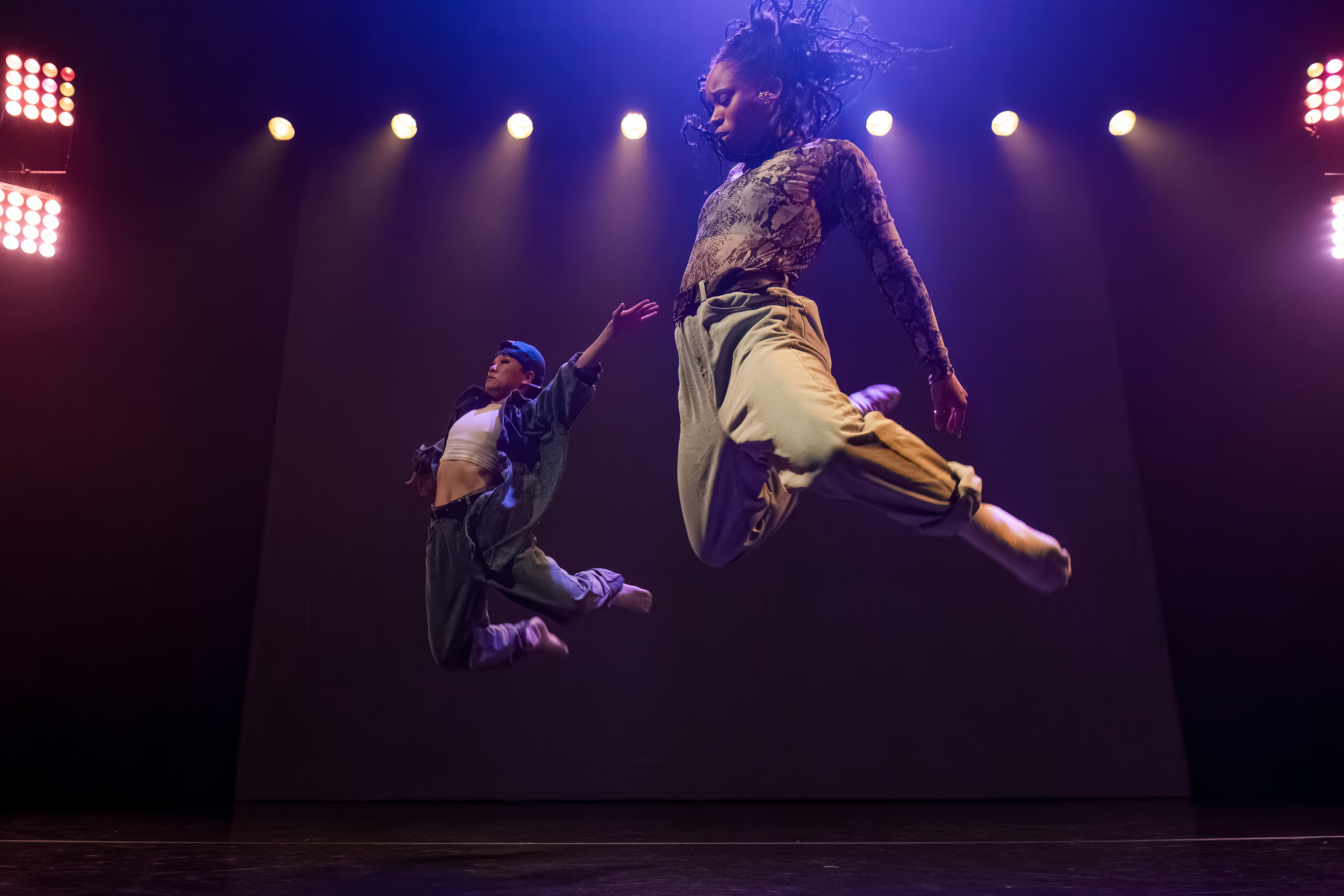 Two dancers leaping in the air lit overhead by purple light and from behind with warm yellow lights.