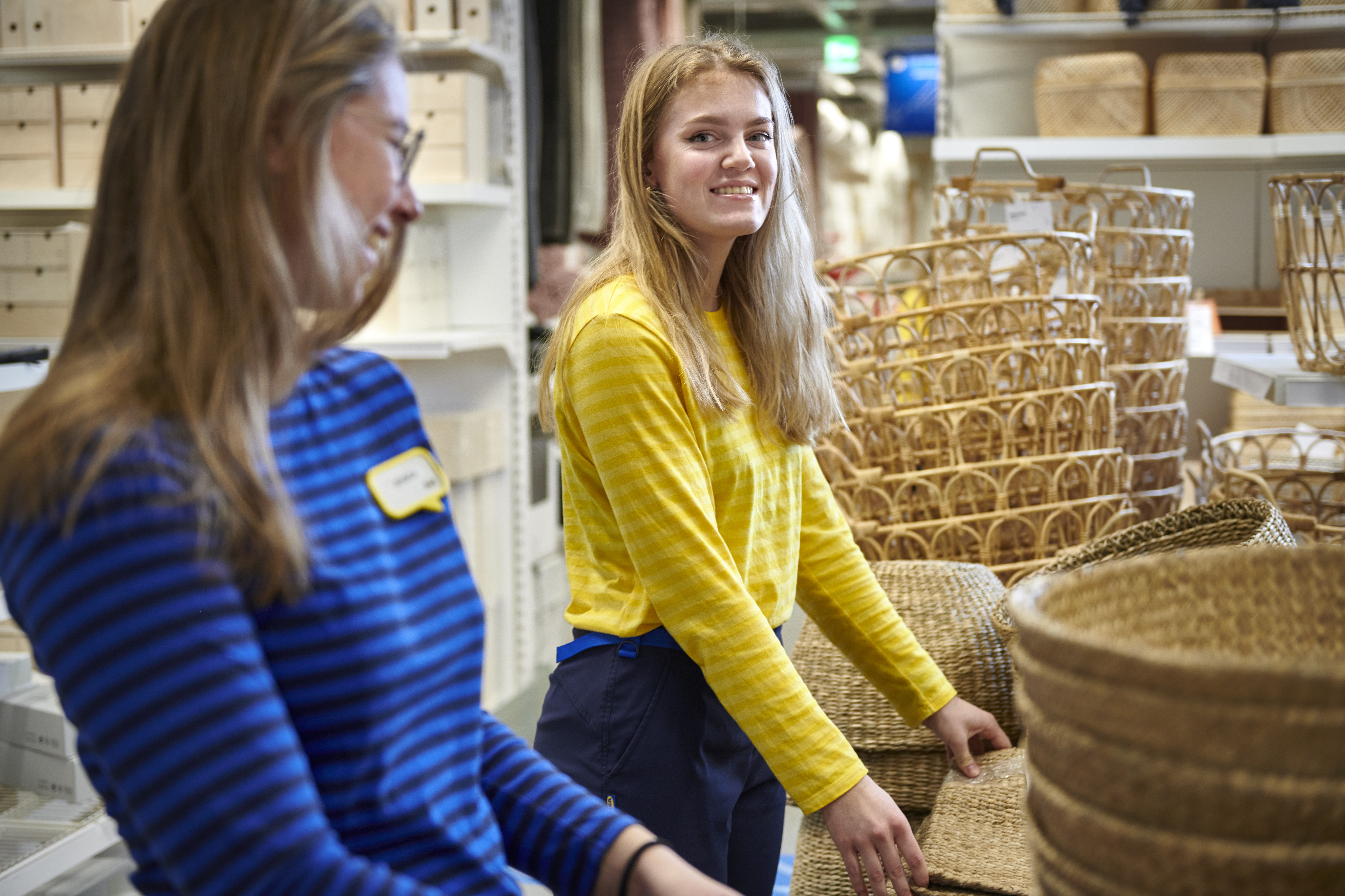 Kaks IKEA töötajat, kellest üks kannab sinise ja teine kollase särgiga vormi, korrastavad poe müügisaalis punutud korvide rida.