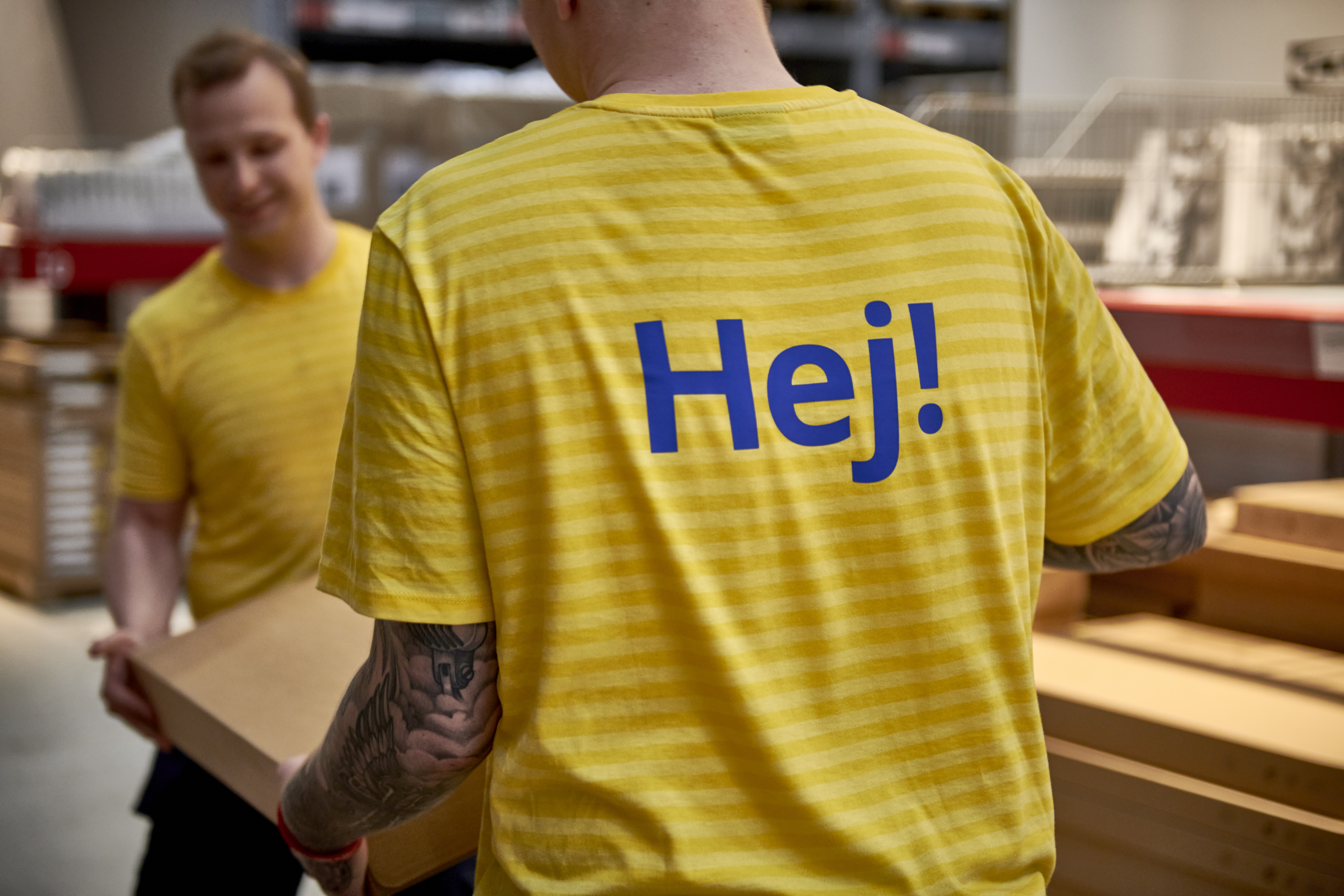 Du IKEA darbuotojai su dryžuotomis geltonos spalvos uniformomis ir užrašu „Hej!“ ant vieno iš jų nugaros kartu neša kartoninę dėžę prekių užsakymo ir atsiėmimo vietoje.