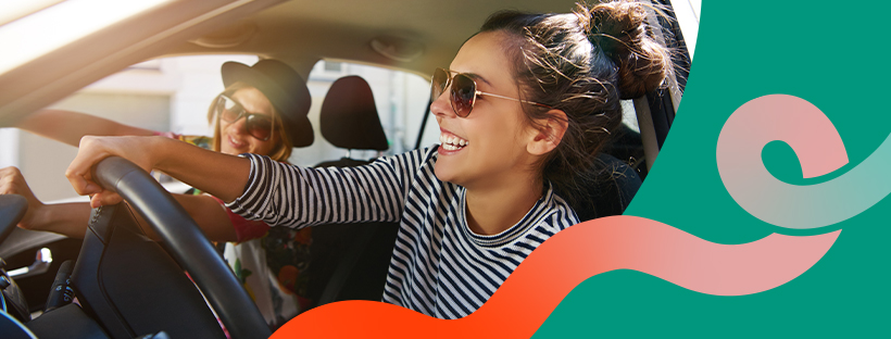 Two smiling women wearing sunglasses enjoying a car ride, one driving and the other in the passenger seat