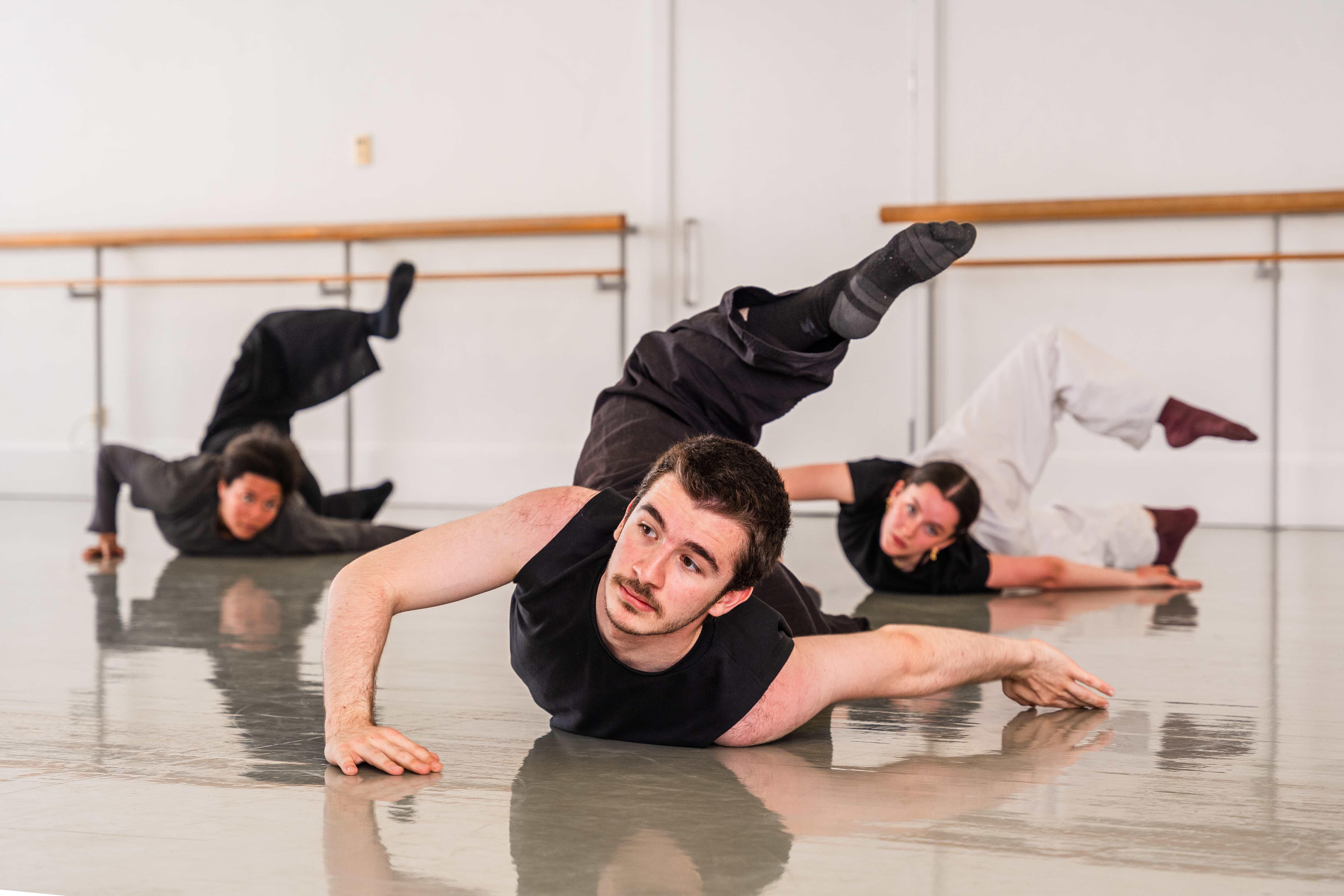 Image of three dancers lying on the floor of a dance studio in a row, their bodies are contorted with one leg in the air and they are looking directly at the camera.
