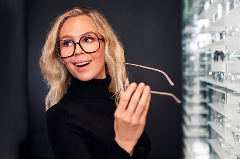 Hymyilevä nainen tummassa poolopaidassa kokeilee silmälaseja Synsam liikkeessä