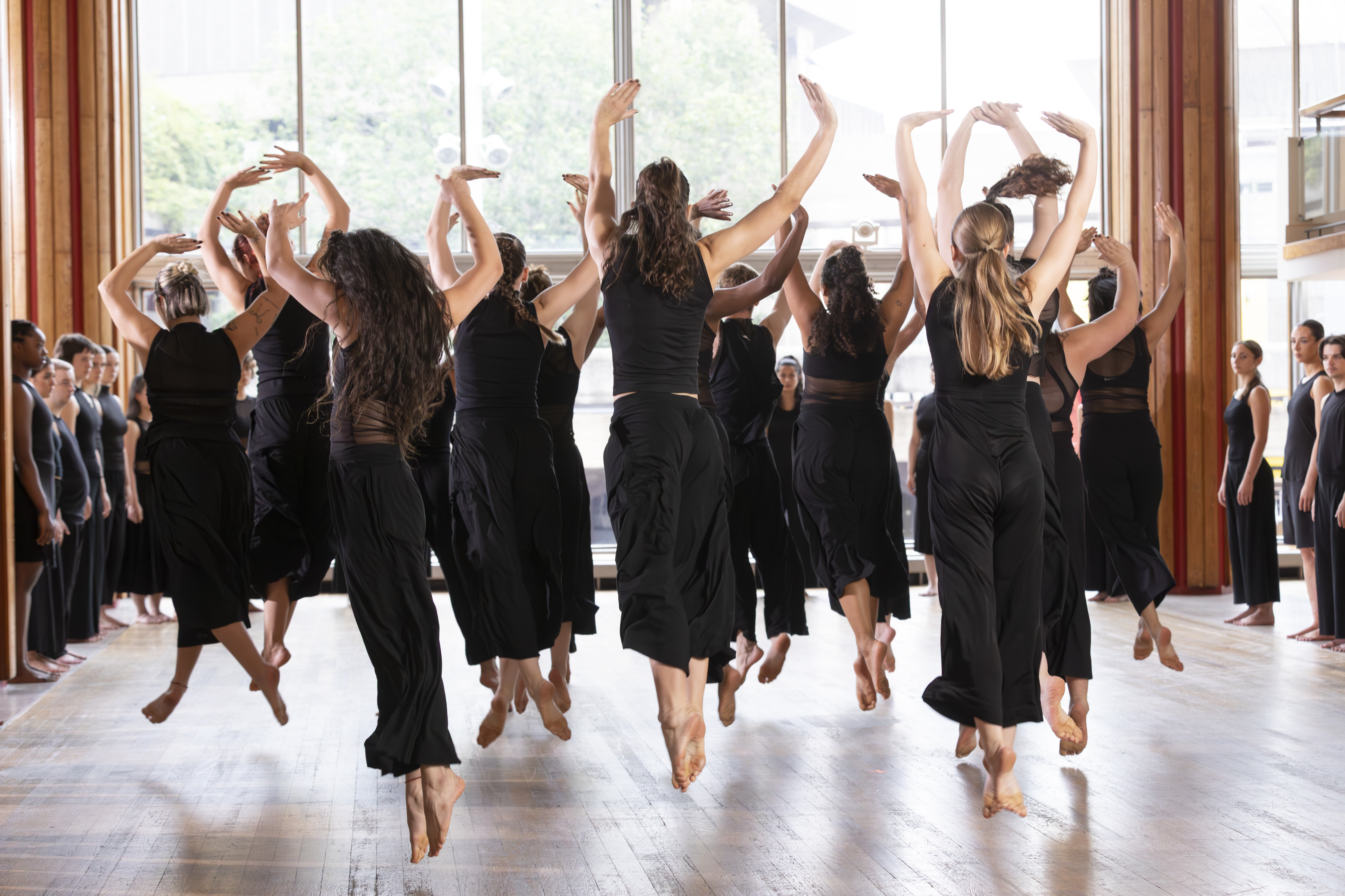 A large group of dancers in black contemporary dance attire perform together in a bright studio with floor-to-ceiling windows. Several dancers are captured mid-jump with their arms raised, while others stand around the edges observing, creating a sense of energy, collaboration, and collective movement.
