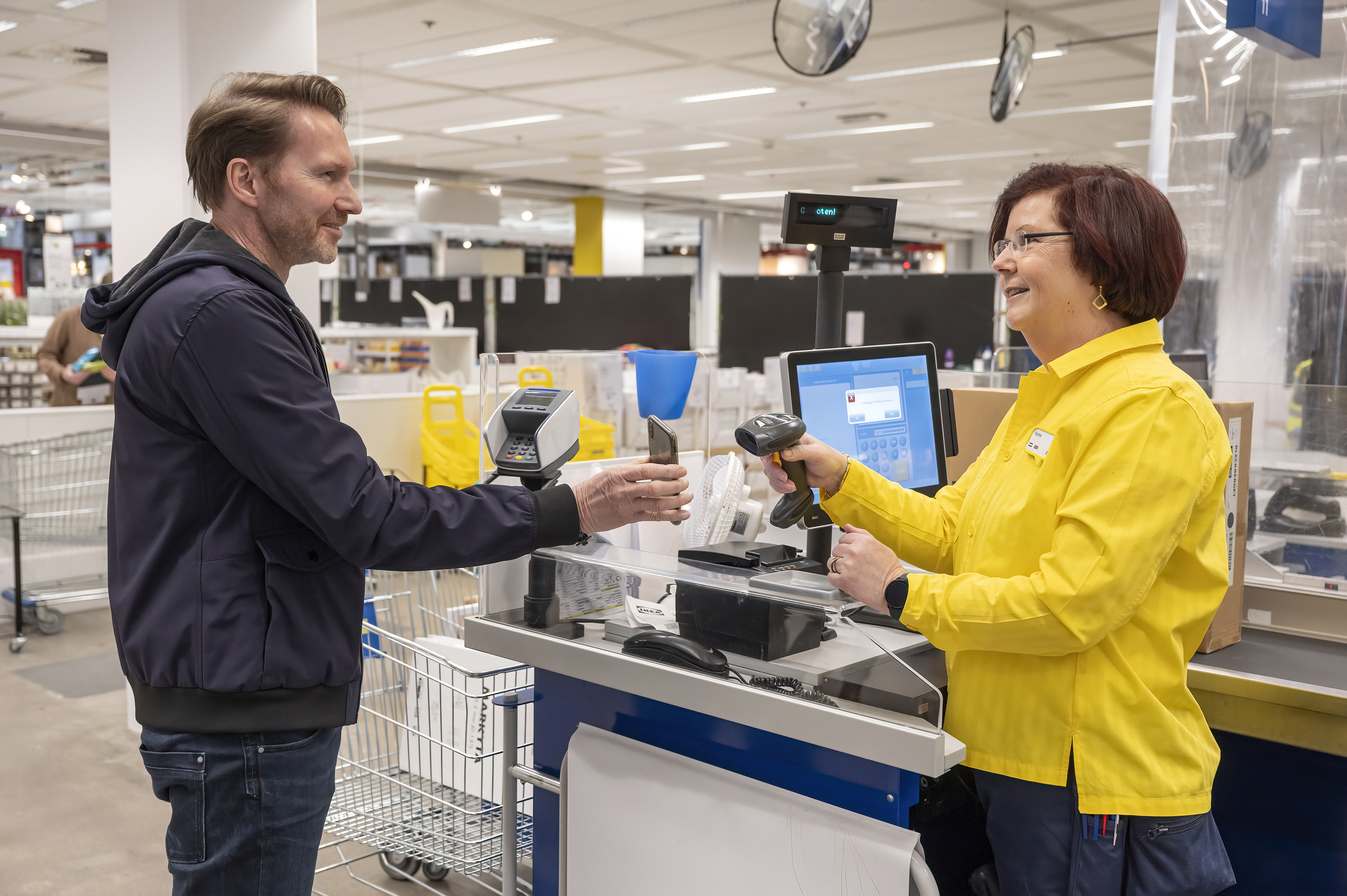 Geltoną uniformą vilkinti IKEA darbuotoja skenuoja prekes prie kasos ir šypsosi pirkėjui, kurį aptarnauja.