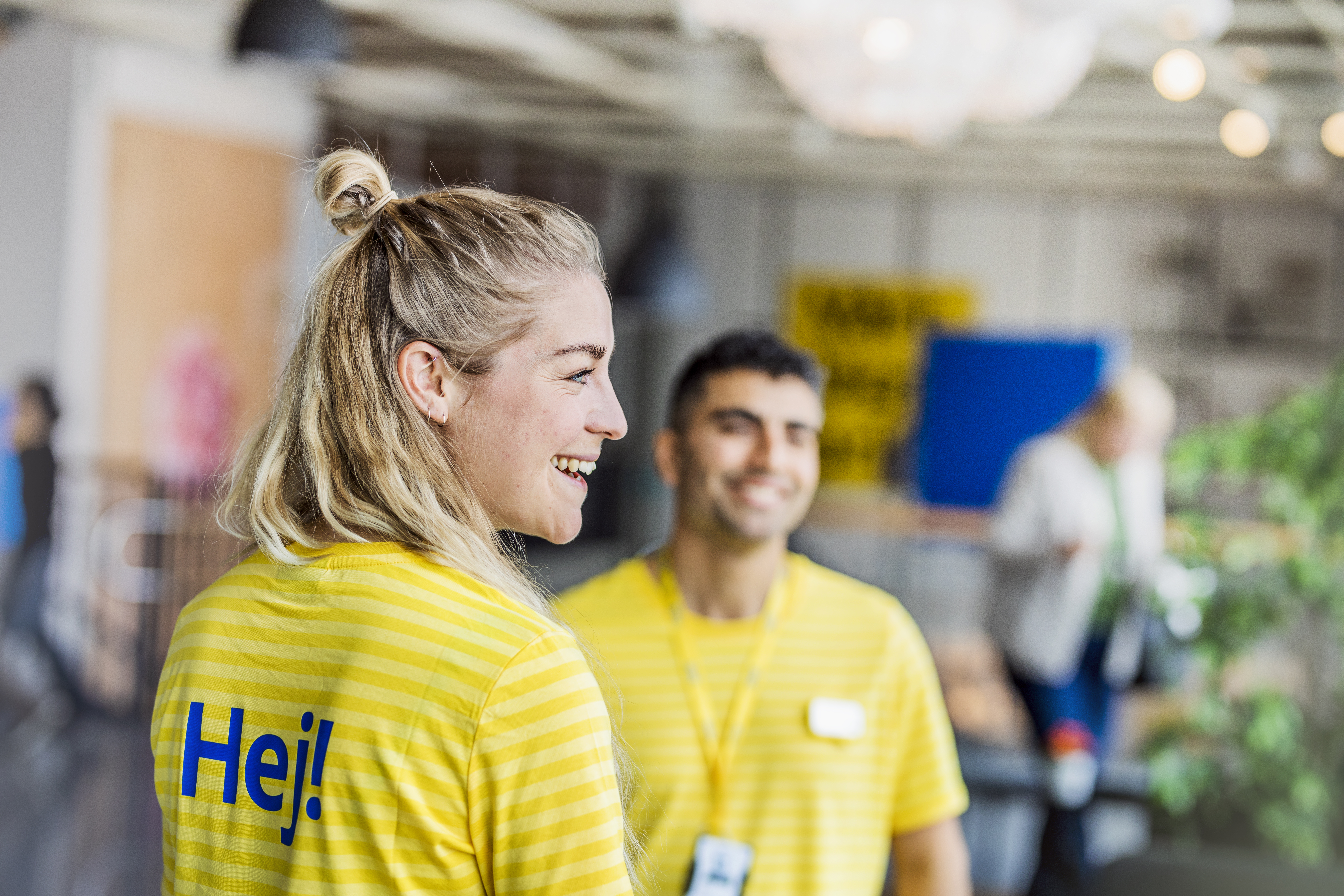 Smaidošs IKEA darbinieks dzeltenā uniformā ar uzrakstu „Hej” uz muguras, fonā redzams vēl viens darbinieks.
