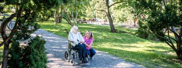 En omsorgsmedarbetare sitter på huk och pratar med en rullstolsburen kund, i en utomhusmiljö