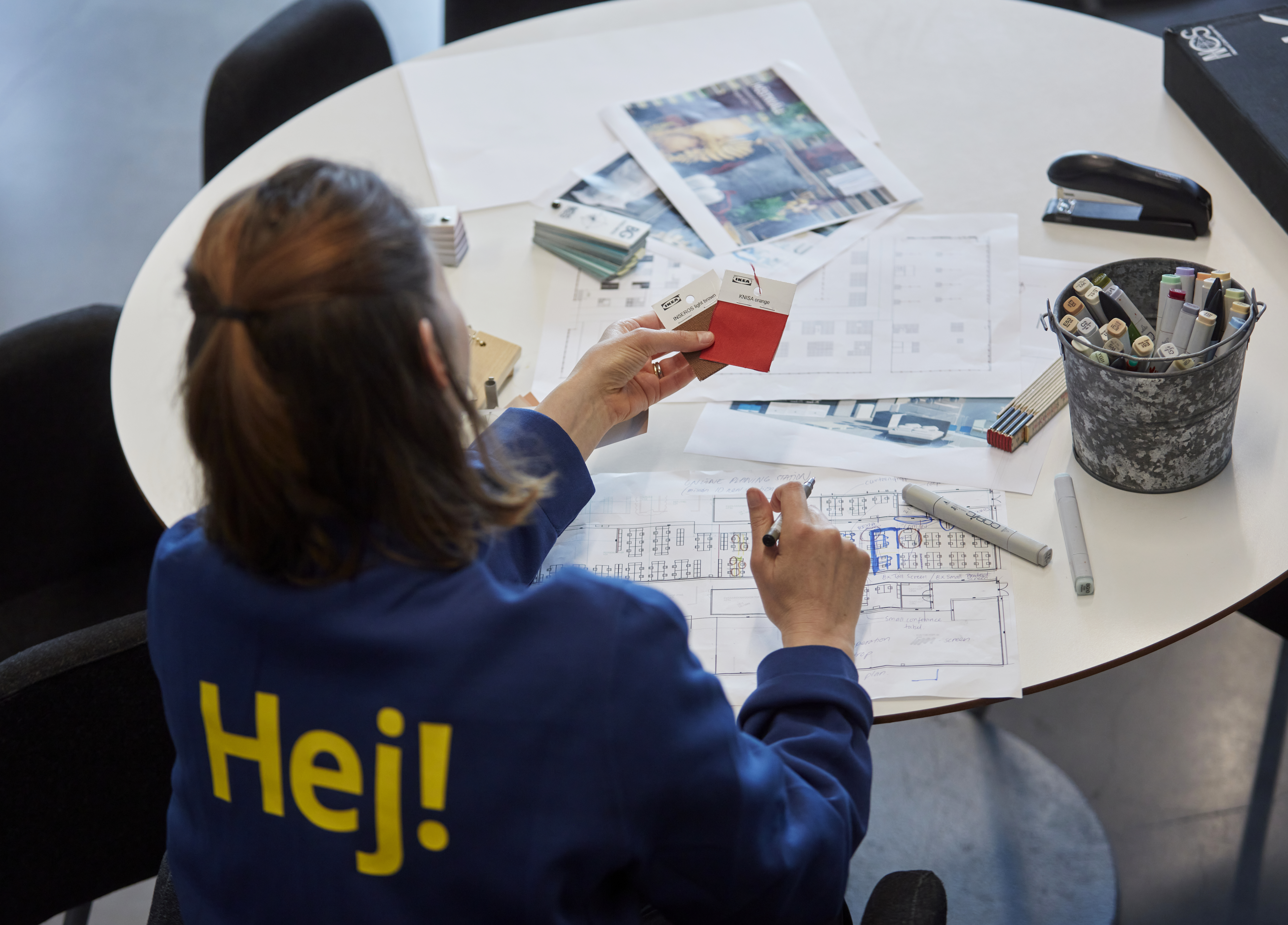 IKEA darbinieks zilā uniformā ar uzrakstu „Hej!” uz muguras sēž pie apaļa galda, pārskata telpu plānojumu un materiālu paraugus, plānojot interjera dizainu.