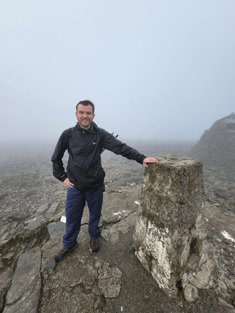 A person in a black waterproof jacket stands beside a stone marker on a foggy mountainside, with rocky terrain and mist obscuring the background. Text overlay reads “Walking with the Ramblers helped me rebuild my life”.
