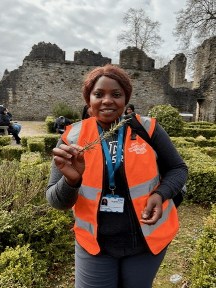 A volunteer wearing a high-visibility orange vest stands in a historic garden holding a sprig of greenery, with stone ruins visible behind. Text overlay reads “Grace’s story: Boosting my community’s health, by volunteering”.