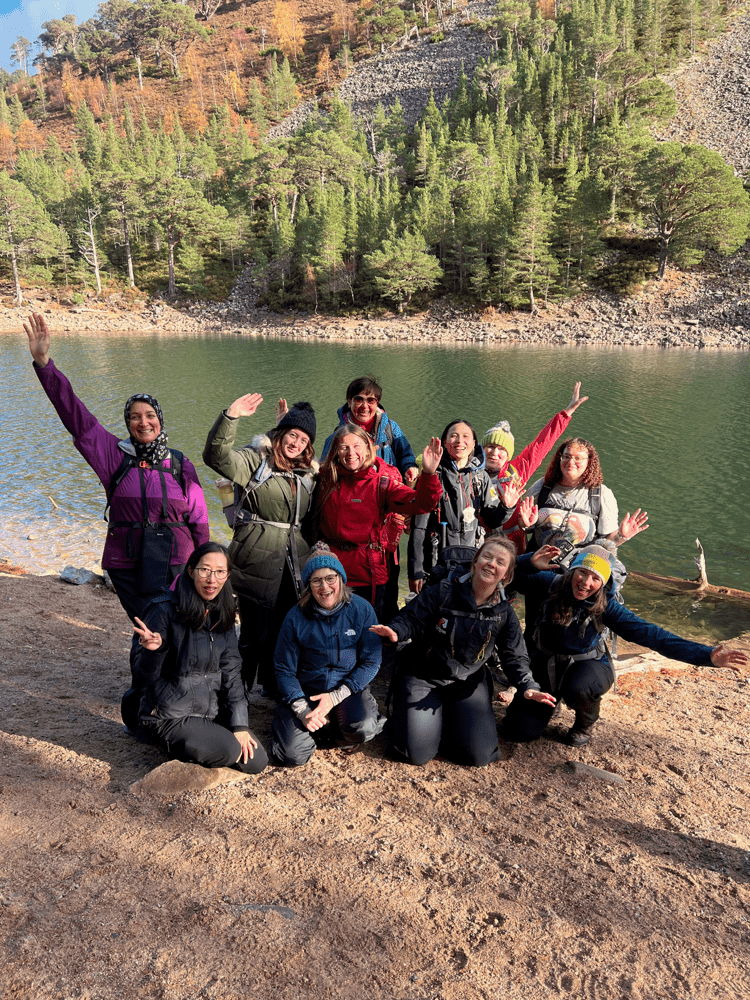 A group of people in outdoor clothing pose beside a lake with trees and hills in the background, some raising their arms. Text overlay reads “Helping young women gain skills and confidence outdoors”.