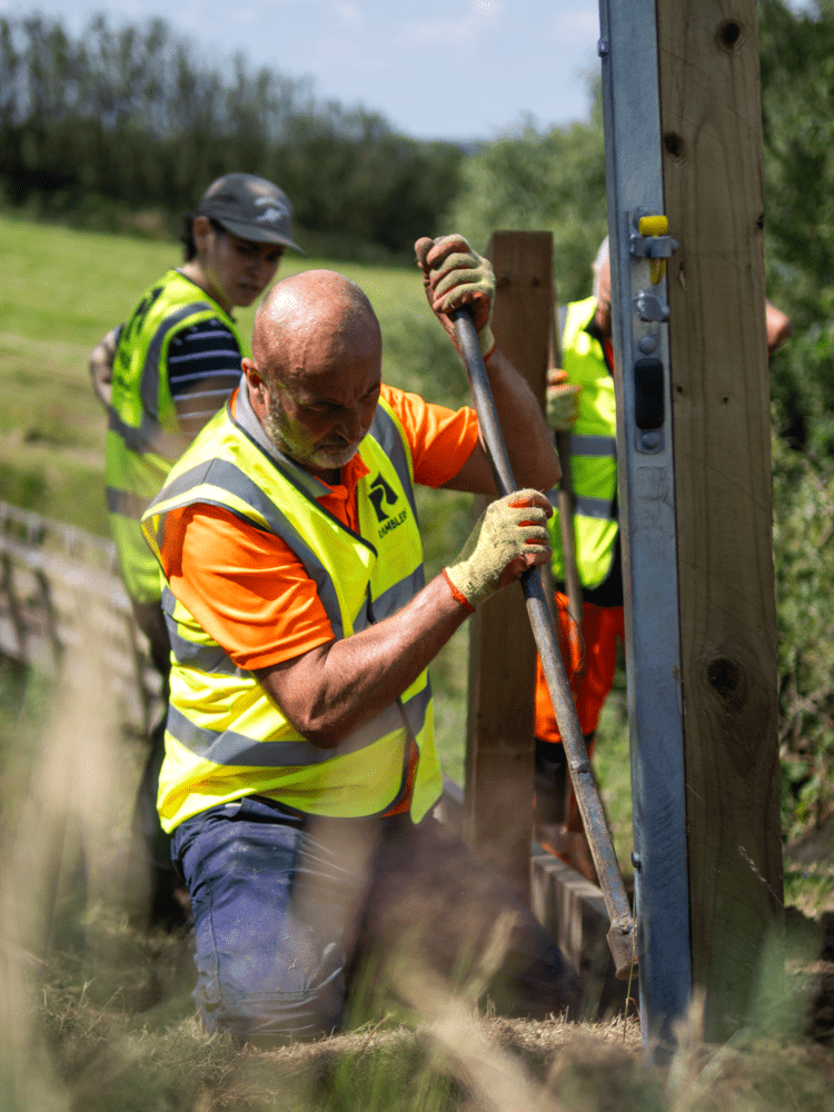 Volunteers wearing high-visibility clothing install a wooden structure alongside a countryside path. Text reads: “Path Maintenance”.